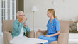 © AboutLife - Nurse in blue uniform gives glass of mineral water to senior lady sitting at small table in light room
