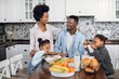 © sofiko14 - Two cute sisters and their young happy parents eating healthy food on bright kitchen during morning time. African american family chatting and smiling during breakfast at cozy home.