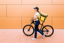 Delivery Woman With Backpack Wheeling Bicycle On Footpath