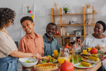 Wall Mural - African american boy looking at mother pouring salad during thanksgiving dinner with grandparents