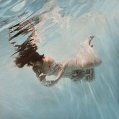 Wall Mural - A girl in a blue dress swims underwater on a light background as if in weightlessness