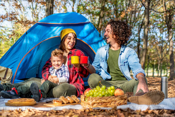 Wall Mural - young happy family camping outdoor with tent in autumn nature. mom, dad and little child having a picnic into the wild. people making a toast with camp mugs enjoying food and drinks. lifestyle concept