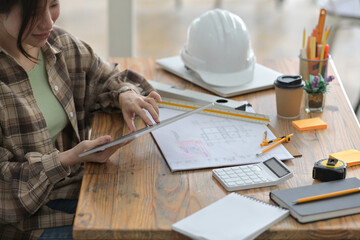 Cropped image of an architect woman's hands is holding a digital tablet at the wooden working desk.