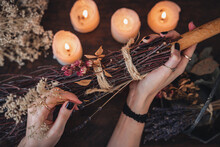 Wiccan Witch Decorating A DIY Besom Broom For Samhain Celebration. Hand Made Broom On A Dark Wooden Table With White Lit Candles And Colorful Dried Flowers And Herbs In The Background