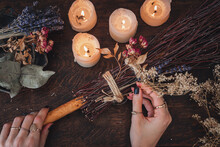Wiccan Witch Decorating A DIY Besom Broom For Samhain Celebration. Hand Made Broom On A Dark Wooden Table With White Lit Candles And Colorful Dried Flowers And Herbs In The Background