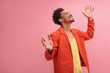 © Look! - Medium shot of happy African young and smiling man with closed eyes with copy space. Well dressed in fall colors with arms raised at his sides against solid pink background.