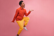 Humorous African Man Sticks Out His Tongue And Raises Leg With Copy Space. Curly Brunette Hair, Yellow Pants And Orange Jacket Make Look Bright, Colorful And Upbeat. Beauty, People Emotions Concept.