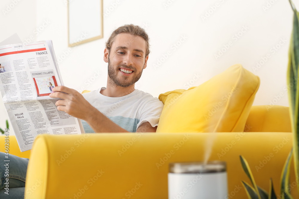 Young man reading newspaper in room with modern humidifier