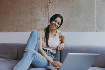 Wall Mural - Happy young latin woman 20s in casual clothes sit on sofa hold use work on laptop pc computer look camera spend time in living room home in own house be lost in reverie good day. Real estate concept