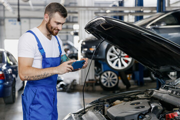 Young professional technician car mechanic man in blue overalls white t-shirt take photo picture on mobile cell phone fix problem with raised hood bonnet work in vehicle repair shop workshop indoor