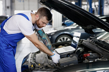 Young repairman professional technician car mechanic man in blue overalls t-shirt using laptop pc computer make diagnostics fix problem with raised hood work in vehicle repair shop workshop indoor