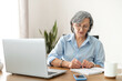© Vadim Pastuh - Serious mature senior female office worker business lady listening to an online webinar on laptop, taking notes in notebook, working on freelance project at home, remote work, learning at home concept