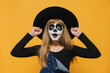 Portrait of shocked excited emotional little child girl with halloween makeup mask open mouth, clenched fists, wear black hat, isolated on yellow color background in studio. Party holiday concept