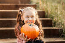 A Little Cheerful Girl Holds An Orange Pumpkin In The Park In The Autumn. Halloween Props