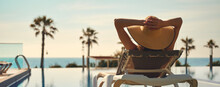 Rear View Woman Wear Hat Lying On Deckchair Near Pool, Put Hands Behind Head Relaxing, Take Sun Bath, Sea Palm Tree Empty Swimming Pool Scenery On Background. Summer Holidays, Vacation, Travel Concept