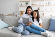 Young asian mother and daughter sitting on sofa and using digital tablet, watching videos or surfing internet
