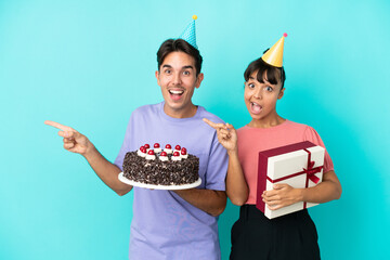 Young mixed race couple holding birthday cake and present isolated on blue background pointing finger to the side and presenting a product