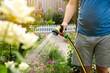 © Andrii Yalanskyi - A gardener with a watering hose and a sprayer water the flowers in the garden on a summer sunny day. Sprinkler hose for irrigation plants. Gardening, growing and flower care concept. Selective focus