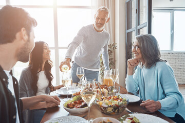 Wall Mural - Happy senior man poring wine and smiling while having dinner with family