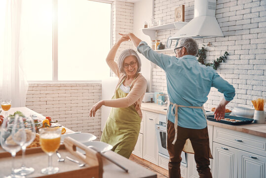beautiful playful senior couple in aprons dancing and smiling while preparing healthy dinner at home