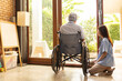 © anon - Attractive young Asian female nurse kneeling beside senior patient in wheelchair talking, smiling and cheering up in comfort at home.He survive from cancer .