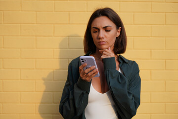 Wall Mural - Caucasian woman wearing shirt at sunset on yellow brick background outdoor positive look at mobile phone screen thoughtful