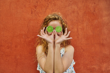 Red haired ukranian woman in casual dress playing with two halfs of lime on orange background