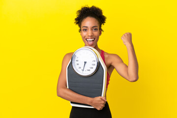 Canvas Print - Young African American woman isolated on yellow background with weighing machine and doing victory gesture