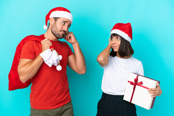 Young couple with christmas hat handing out gifts isolated on blue background having doubts while scratching head
