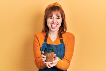 Poster - Redhead young woman wearing waitress apron holding take away cup of coffee sticking tongue out happy with funny expression.