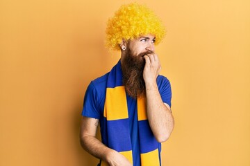 Wall Mural - Redhead man with long beard football hooligan cheering game wearing funny wig looking stressed and nervous with hands on mouth biting nails. anxiety problem.