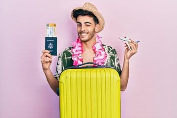 Wall Mural - Young hispanic man wearing summer style and hawaiian lei holding passport and plane toy winking looking at the camera with sexy expression, cheerful and happy face.