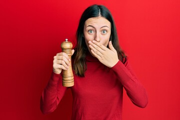 Young brunette woman black pepper grinder covering mouth with hand, shocked and afraid for mistake. surprised expression