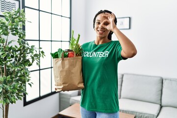 Sticker - Young african american woman wearing volunteer t shirt holding bag of groceries smiling happy doing ok sign with hand on eye looking through fingers