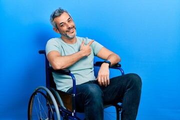 Canvas Print - Handsome middle age man with grey hair sitting on wheelchair cheerful with a smile on face pointing with hand and finger up to the side with happy and natural expression