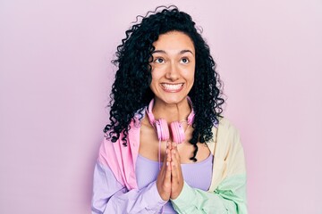 Young hispanic woman with curly hair wearing gym clothes and using headphones begging and praying with hands together with hope expression on face very emotional and worried. begging.