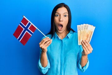 Wall Mural - Young latin woman holding norway flag and krone banknotes afraid and shocked with surprise and amazed expression, fear and excited face.