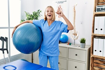 Wall Mural - Young caucasian physiotherapist woman holding pilates ball surprised with an idea or question pointing finger with happy face, number one