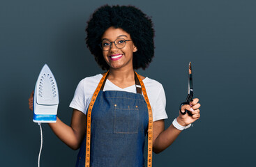 Canvas Print - Young african american woman dressmaker designer wearing atelier apron holding iron and scissors smiling with a happy and cool smile on face. showing teeth.