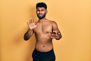 Poster - Arab man with beard wearing swimwear shirtless afraid and terrified with fear expression stop gesture with hands, shouting in shock. panic concept.