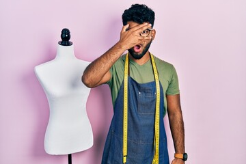 Canvas Print - Arab man with beard dressmaker designer wearing atelier apron peeking in shock covering face and eyes with hand, looking through fingers afraid