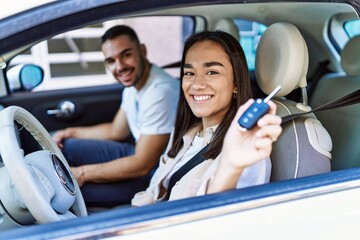 Young hispanic couple driving auto at the city. Girl smiling happy holding key of new car.