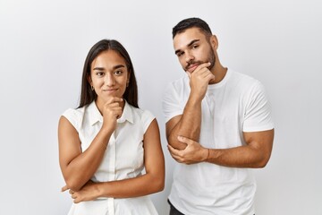 Poster - Young interracial couple standing together in love over isolated background looking confident at the camera with smile with crossed arms and hand raised on chin. thinking positive.