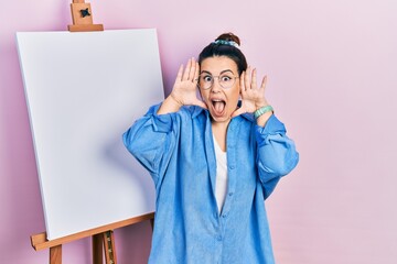 Poster - Young hispanic woman standing by painter easel stand smiling cheerful playing peek a boo with hands showing face. surprised and exited