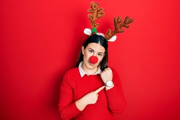 Canvas Print - Young hispanic woman wearing deer christmas hat and red nose in hurry pointing to watch time, impatience, looking at the camera with relaxed expression