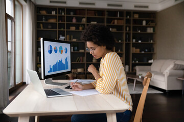 Concentrated young african american businesswoman employee worker in eyeglasses analyzing online sales statistics data on computer, reviewing marketing research, working with documents at home office.