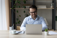Smiling Confident Businessman In Glasses Working On Laptop Online, Sitting At Home Office Desk, Looking At Camera, Happy Successful Man Entrepreneur Freelancer Or Student Typing, Writing Message