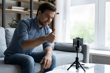 Sticker - Smiling young man showing thumb up, using smartphone on tripod, satisfied blogger influencer recommending service, recording video for social network on mobile device, sitting on couch at home