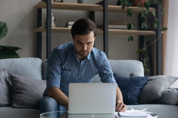 Poster - Serious man using laptop, working or studying online, sitting on couch at home, focused confident businessman freelancer or student looking at computer screen, watching webinar, taking notes