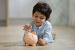 © fizkes - Happy small adorable mixed race child boy putting coins in piggybank, learning saving money for future purchase, calculating personal budget or managing finances, lying on warm heated floor.